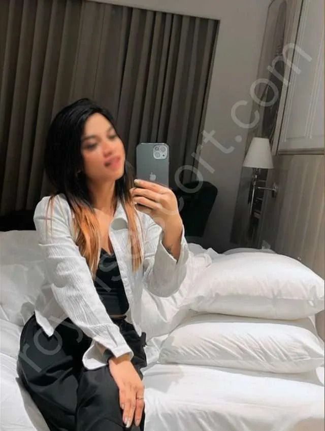 Naira escort in Zirakpur wearing black dress with white shirt and sitting on bed taking selfii 
